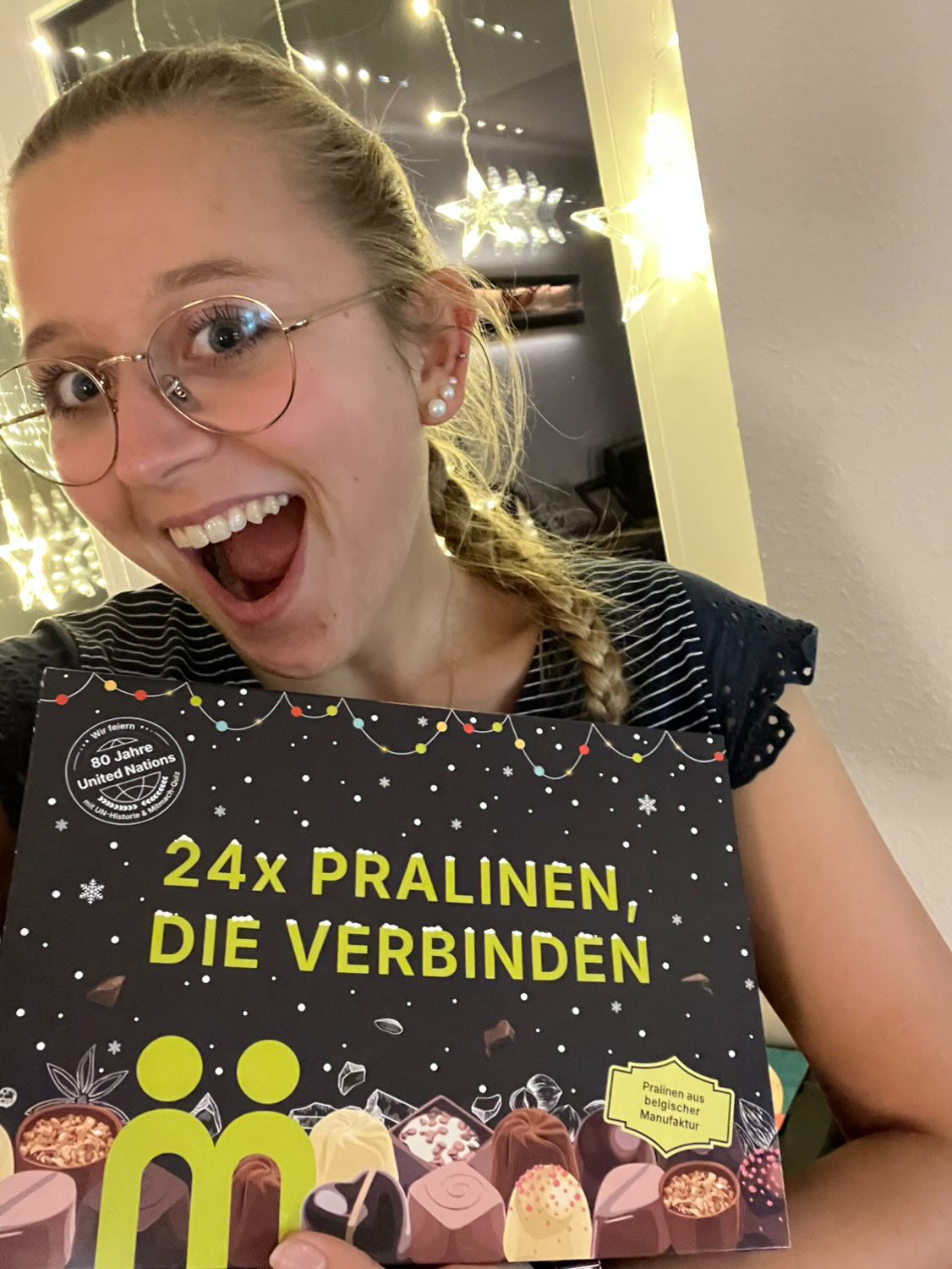 myBuddy Community-Foto: Junge Frau mit Brille, Mund offen vor Freude, hält den PRALINEN-Kalender vor Lichterketten