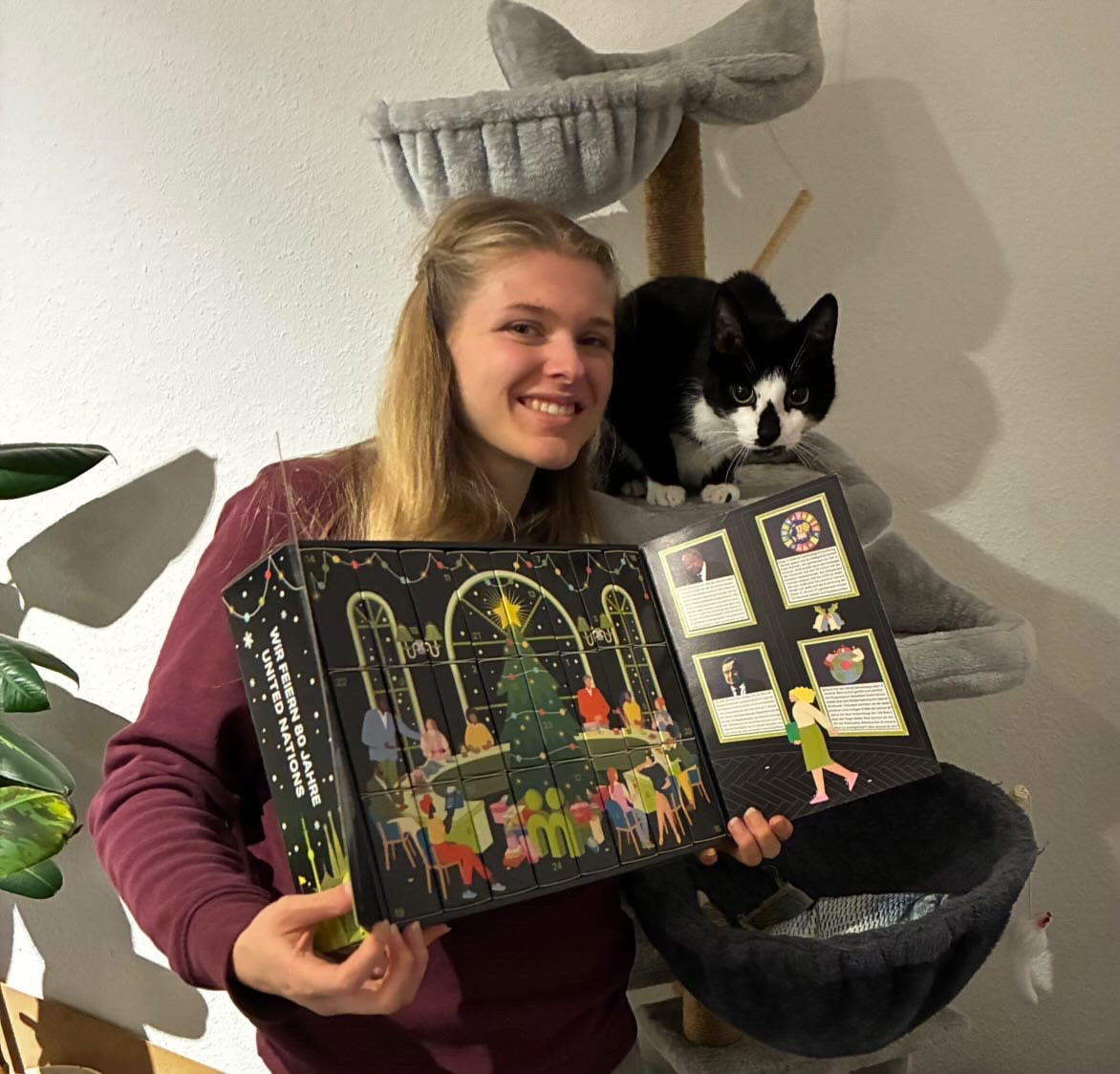 myBuddy Community-Foto: Frau im weinroten Pulli zeigt den aufgeklappten Kalender, schwarz-weiße Katze auf dem Kratzbaum dahinter