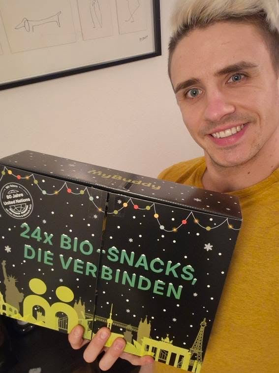 myBuddy Community-Foto: Mann im gelben Shirt hält den Kalender ins Selfie, gerahmte Strichzeichnungen an der Wand