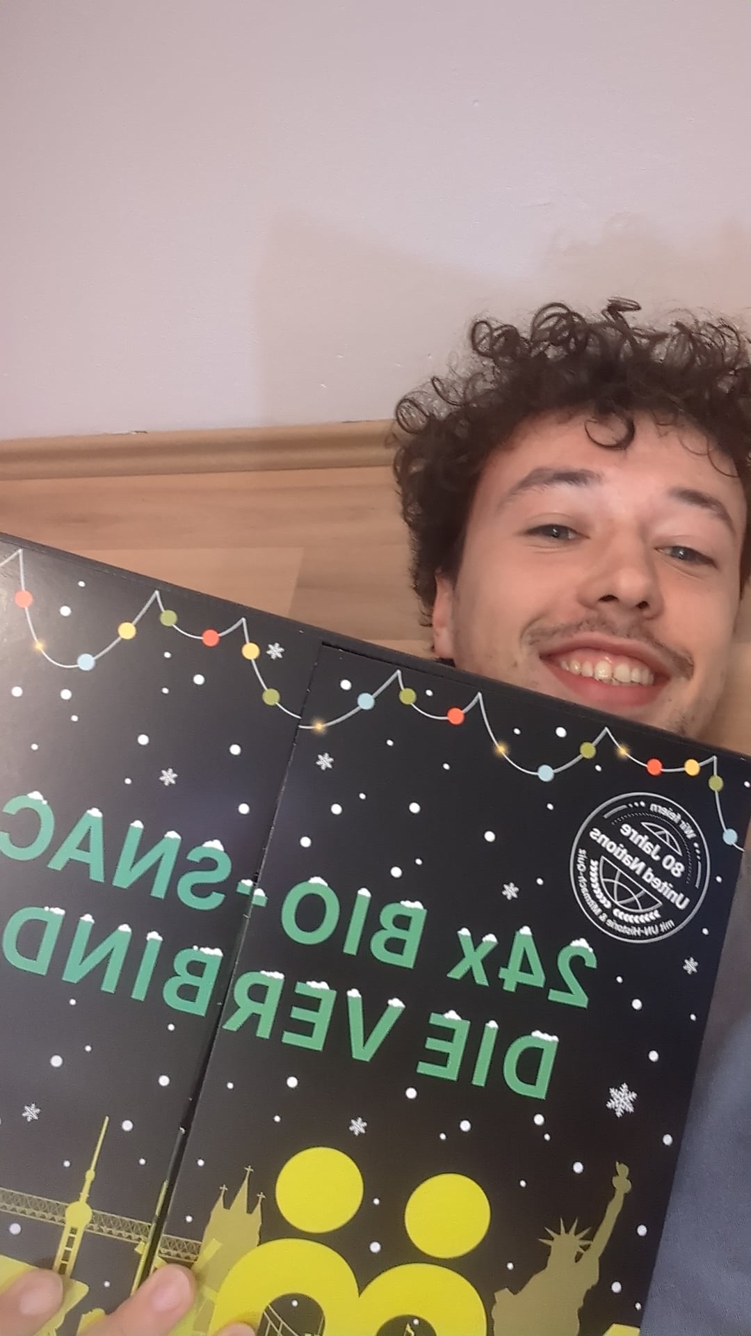 myBuddy Community-Foto: Junger Mann mit Lockenkopf hält den Kalender diagonal ins Selfie, heller Flur