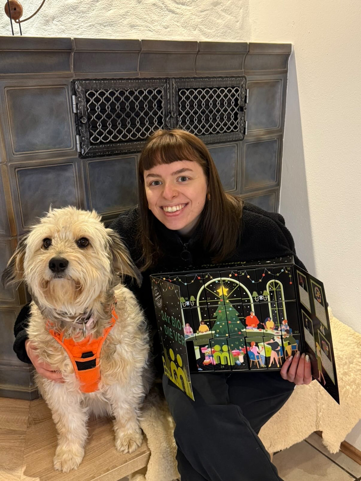 myBuddy Community-Foto: Frau mit Pony sitzt vor dem Kachelofen, Arm um einen hellen Terrier, Kalender aufgeklappt