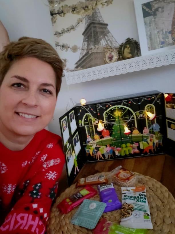 myBuddy Community-Foto: Frau im Weihnachtspulli neben dem beleuchteten aufgeklappten Kalender, Türchen-Inhalte auf dem Tisch ausgebreitet