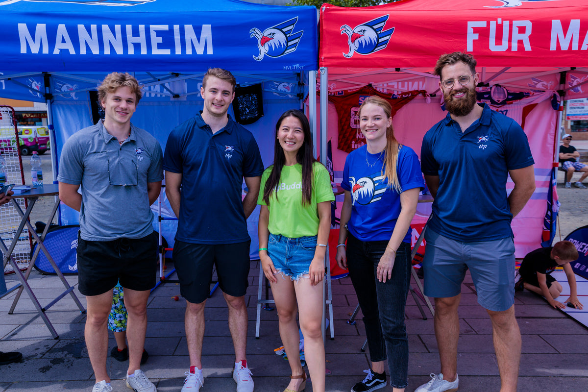 myBuddy Über uns: Fünf Personen stehen nebeneinander vor einem blauen und einem roten Pavillon mit Adler-Logo und dem Schriftzug „Mannheim“, in der Mitte eine Frau im grünen myBuddy-Shirt
