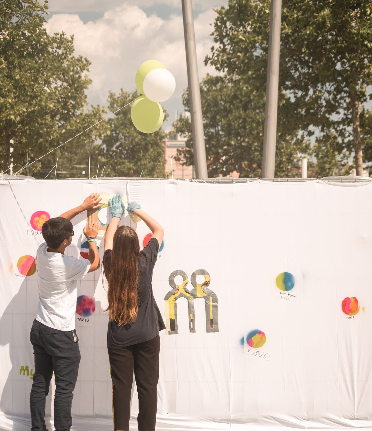 myBuddy Über uns: Zwei Jugendliche kleben von hinten ein großes myBuddy-Logo an eine weiße Stoffwand voller bunter Farbkleckse, daneben steigen grüne und weiße Luftballons auf