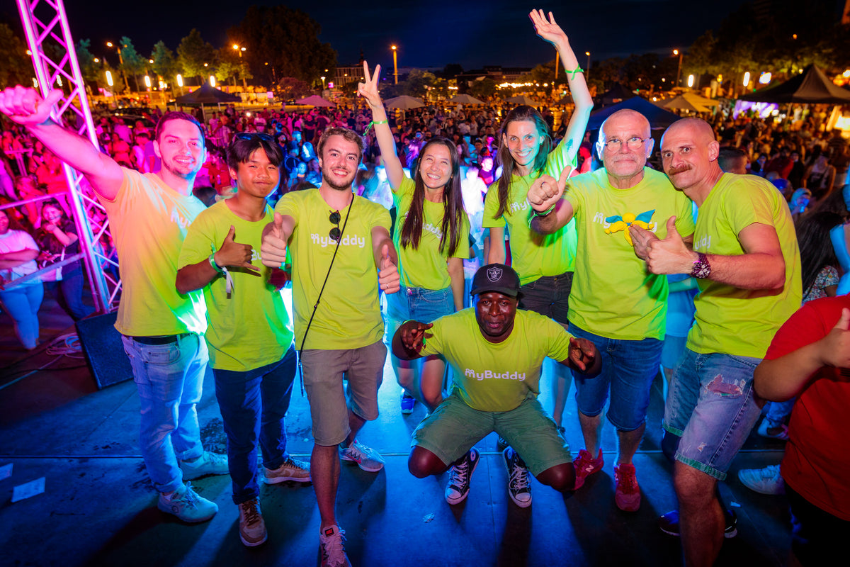 myBuddy Über uns: Acht Personen in neongrünen myBuddy-Shirts jubeln auf einer Festivalbühne bei Nacht, davor kniet ein Mann mit Cap, dahinter das Publikum