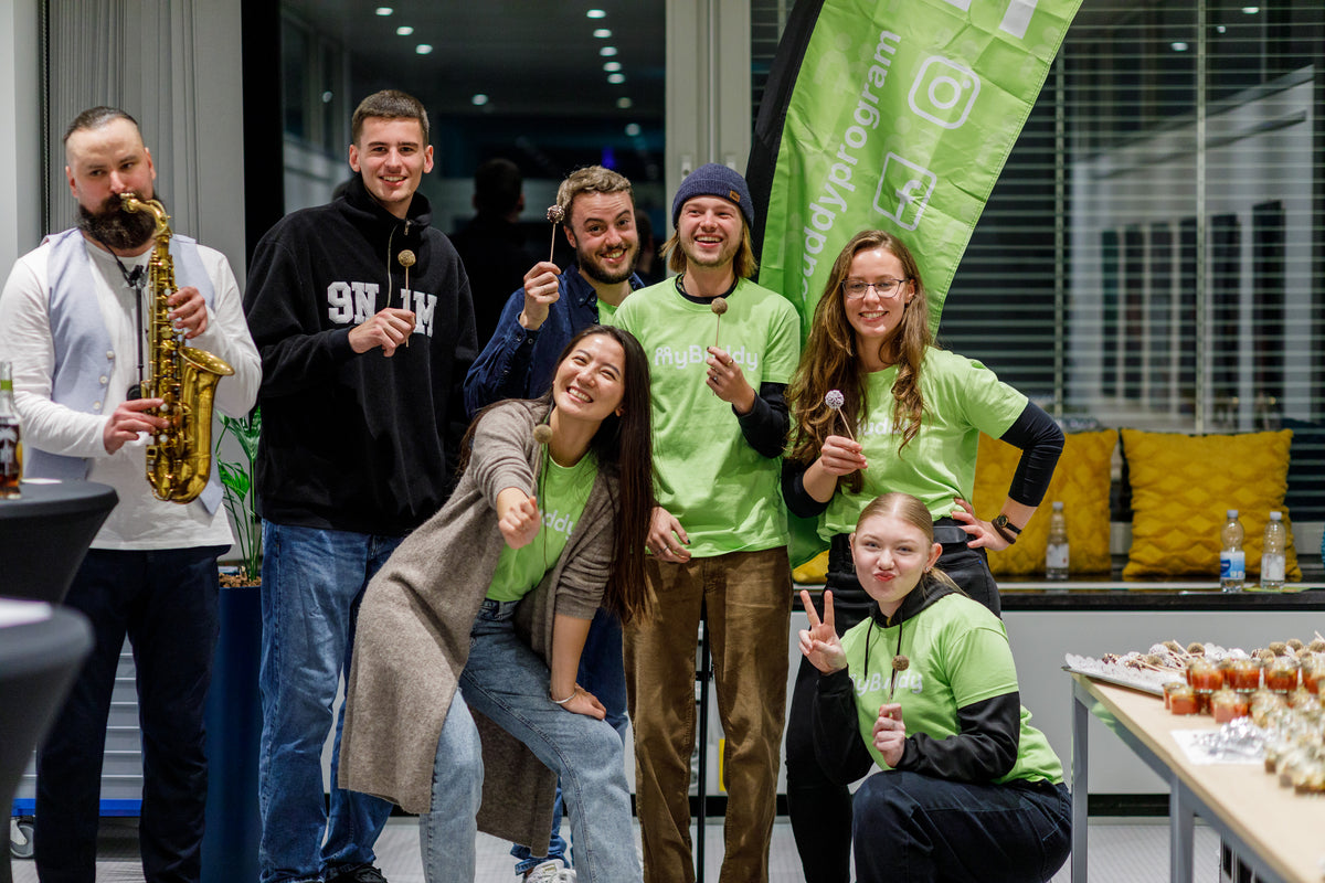 myBuddy Über uns: Gruppe in grünen myBuddy-Shirts hält Cakepops in die Kamera, links spielt ein Mann Saxofon, dahinter eine grüne Beachflag mit Instagram-Logo
