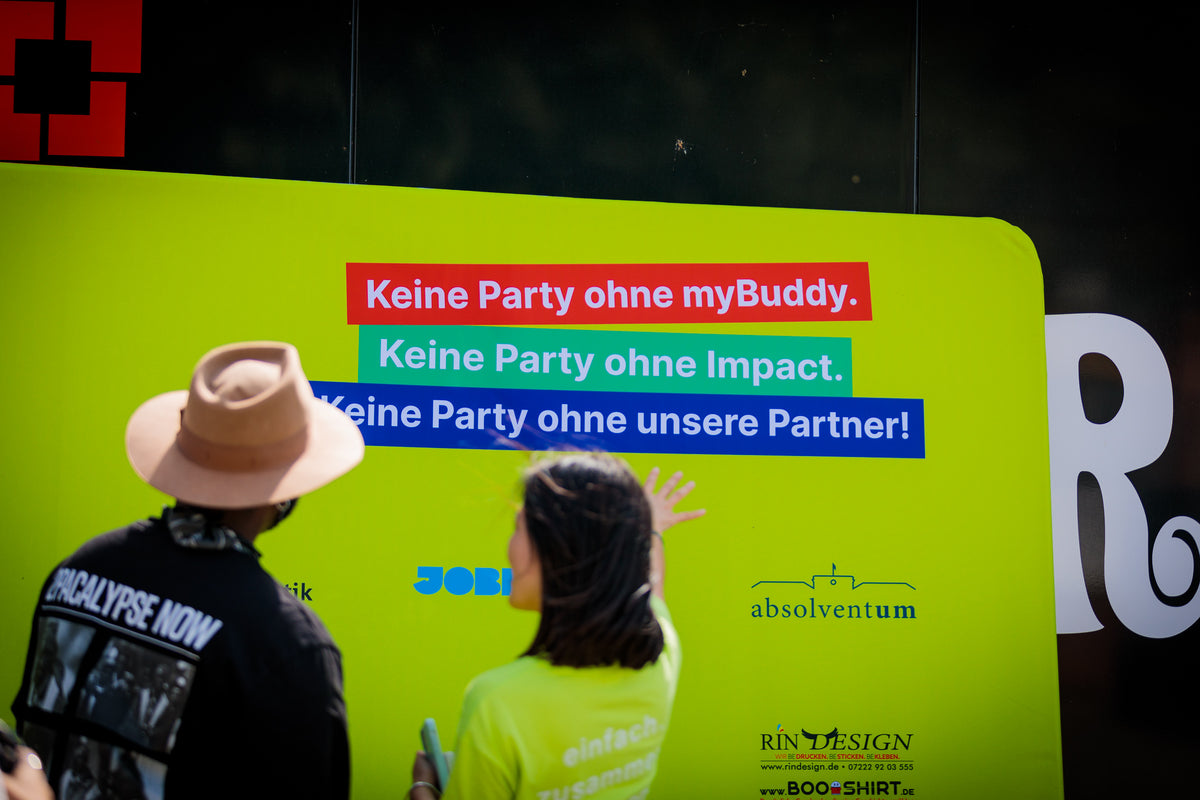 myBuddy Über uns: Zwei Personen von hinten vor einer grünen Partnerwand mit dem Text „Keine Party ohne myBuddy. Keine Party ohne Impact. Keine Party ohne unsere Partner!“