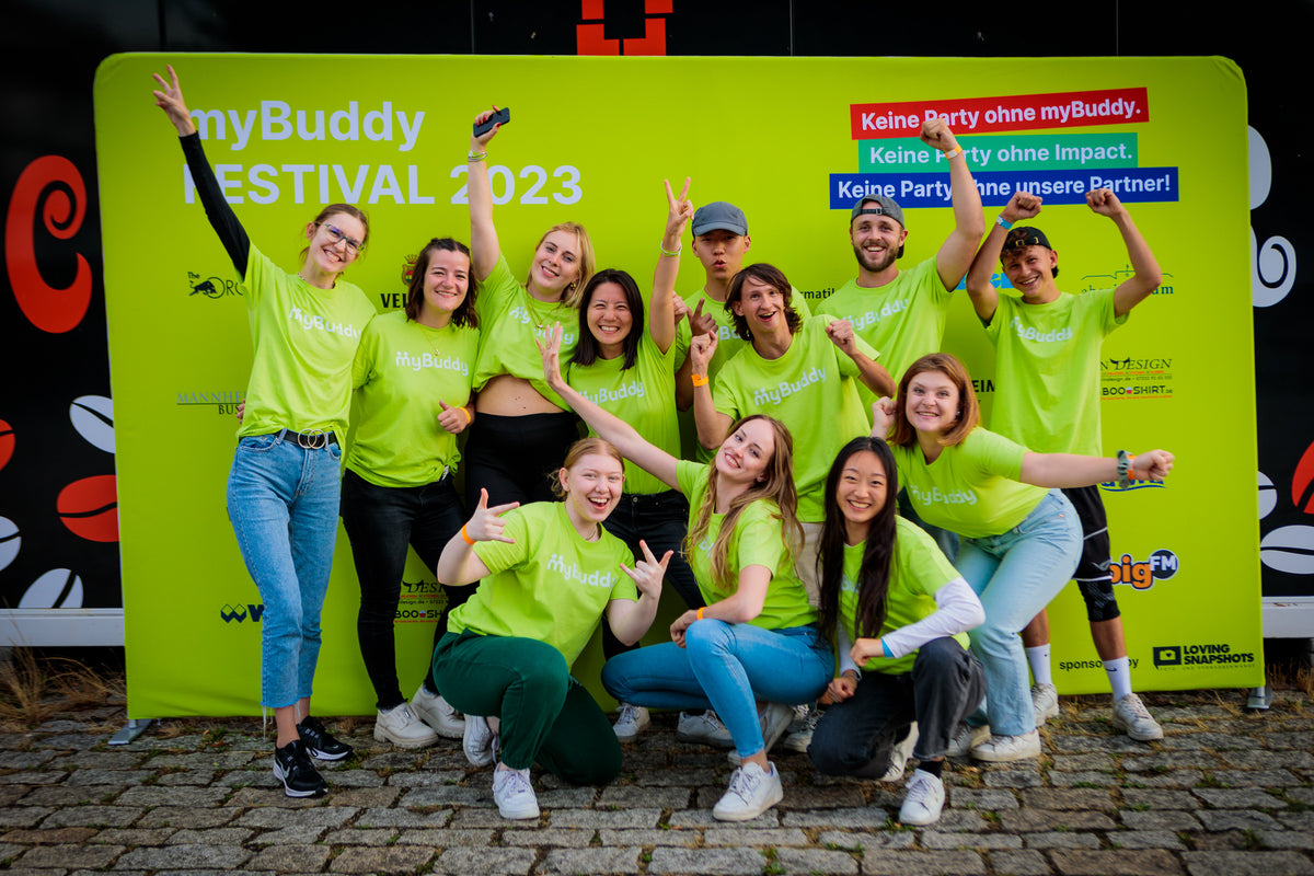 myBuddy Über uns: Zwölf Personen in neongrünen myBuddy-Shirts posieren mit erhobenen Armen vor einer grünen Fotowand mit dem Schriftzug „myBuddy FESTIVAL 2023“