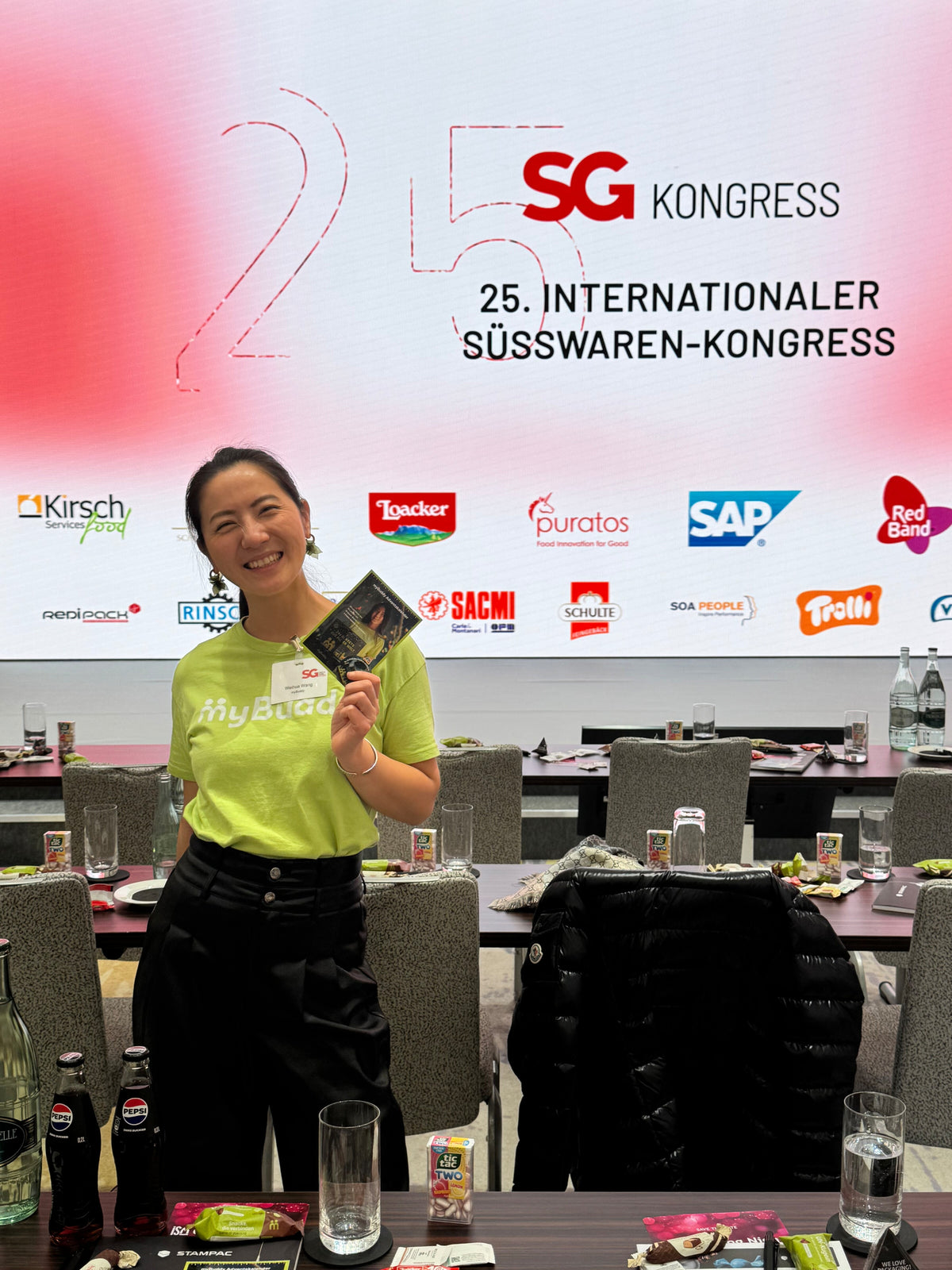 myBuddy Über uns: Frau im grünen myBuddy-Shirt hält einen myBuddy-Adventskalender-Flyer hoch, dahinter eine Leinwand „25. Internationaler Süsswaren-Kongress“ mit Sponsorenlogos